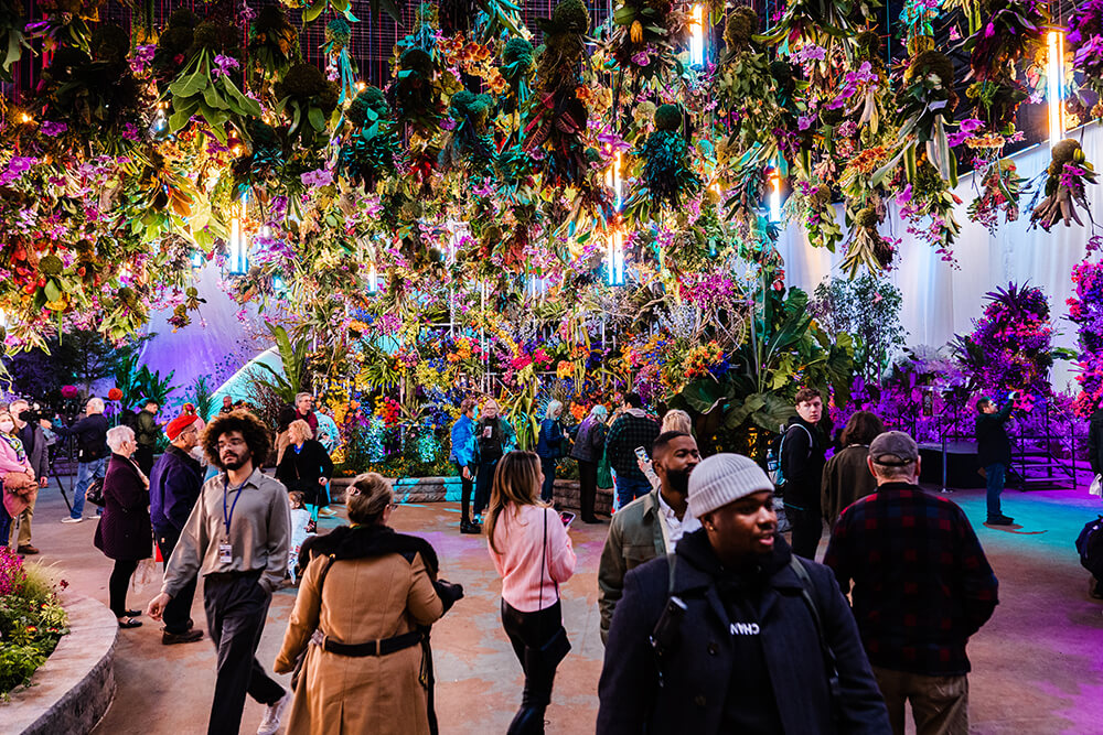 Bright, colorful floral arrangements hang from the ceiling. Flower Show attendees walk underneath, marveling at the floral displays surrounding them. A large floral display is in the center with a crowd of people admiring it.