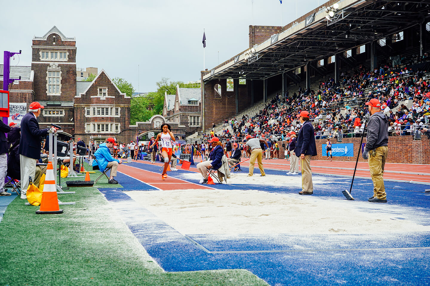 Runner approaching long jump pit of sand as officials stand nearby monitoring event, crowded stands packed with fans behind