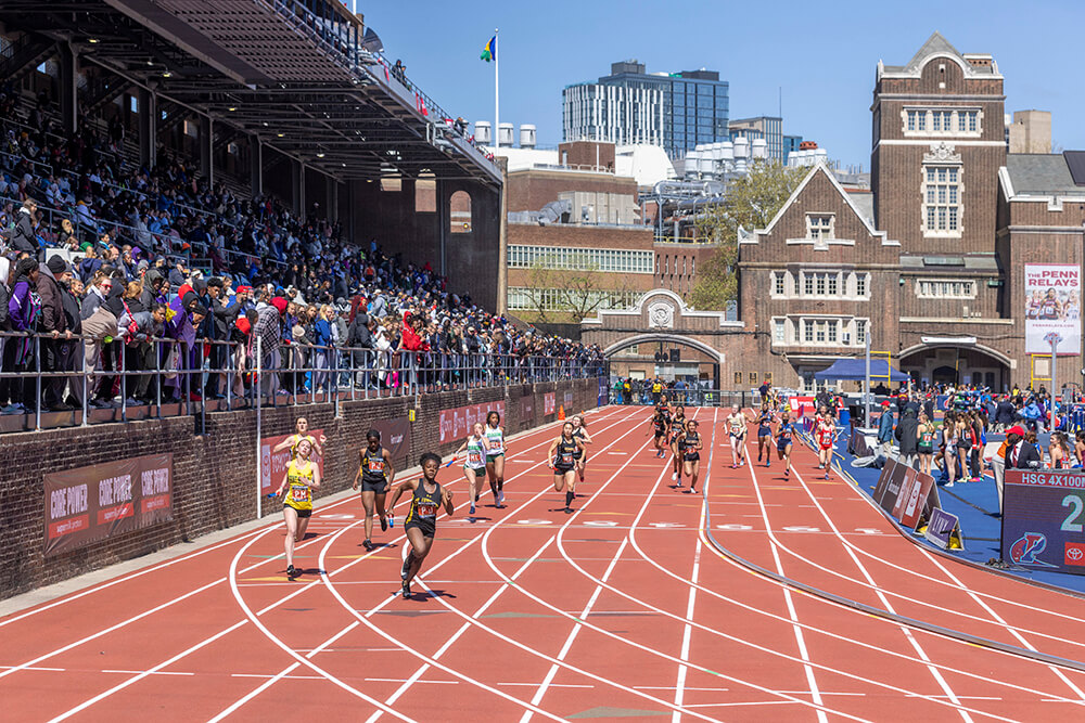 Runners are rounding a track as they run with batons in their hands. A crowd cheers from the bleachers to the left. Officials and coaches stand to the right. The campus is shown in the background.