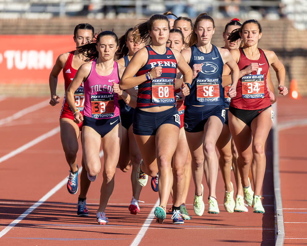 Several female athletes are running on a track. They are running toward the camera. Each one is wearing a number on her school's jersey.