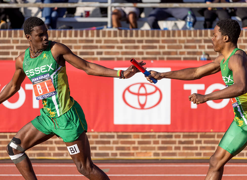 Two men in green uniforms are shown competing in a relay race. One male athlete is shown passing the baton to his teammate.