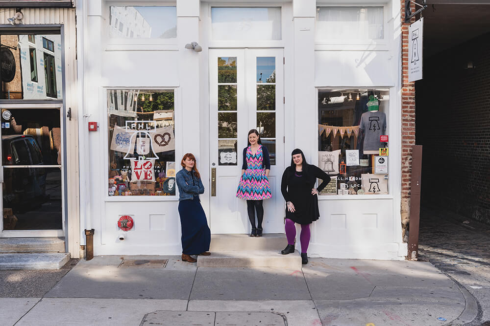 Three women stand in front of a storefront. The woman to the left of the front door has reddish hair and is wearing a jean jacket over a long navy dress with brown boots. The woman in the middle has dark hair and is wearing a dark short sweater over a brightly colored pink and blue dress with black stockings and shoes on. The woman to the right also has dark hair. She wears a black dress, purple leggings and black shoes. The windows of the storefront show Philadelphia-themed gifts and souvenirs for sale.