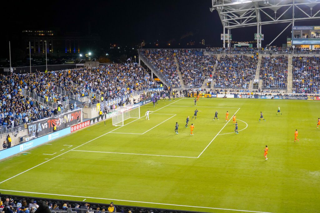 A soccer field with two teams playing, one with its players dressed in orange uniforms, the other team's players are dressed in blue uniforms. The stands are filled with fans attentively watching the playoff game