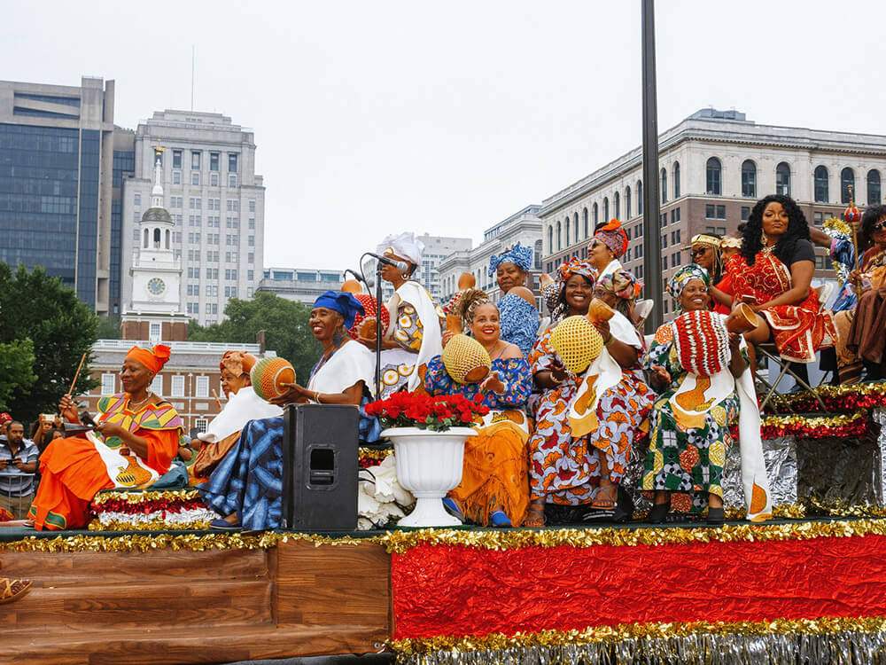 More than a dozen women dressed in bright outfits and wearing beautiful headdresses are shown sitting on a colorful float during a Juneteenth parade. Independence Hall is in the background.