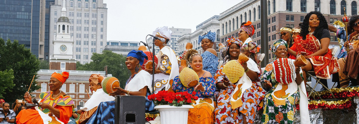 More than a dozen women dressed in bright outfits and wearing beautiful headdresses are shown sitting on a colorful float during a Juneteenth parade. Independence Hall is in the background.