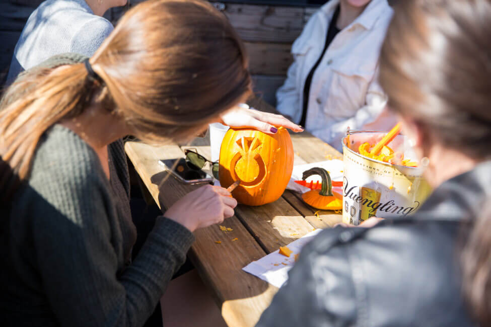 A woman is shown carving a ring into a pumpkin on a picnic table. Her phone is on the table to the left of the pumpkin. A bucket is off to the right of the pumpkin.