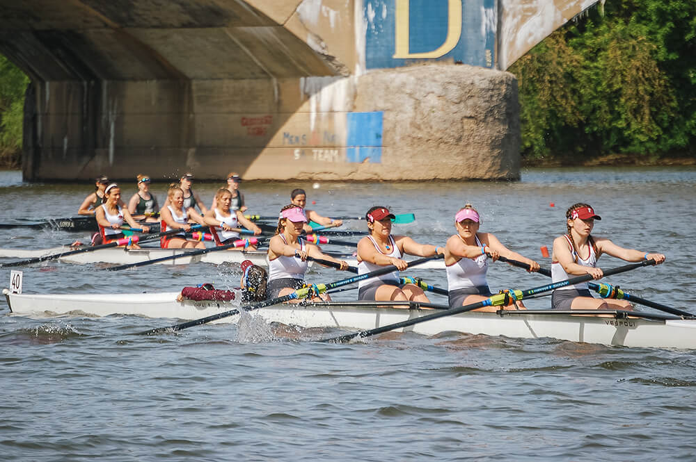 Three boats of four rowers are shown in the river during the Dad Vail Regatta. The four women in the boat closest to the camera are wearing visors, the first and third women are wearing red visors, the second and fourth women are wearing pink visors. Water splashes up from their oars in the river.