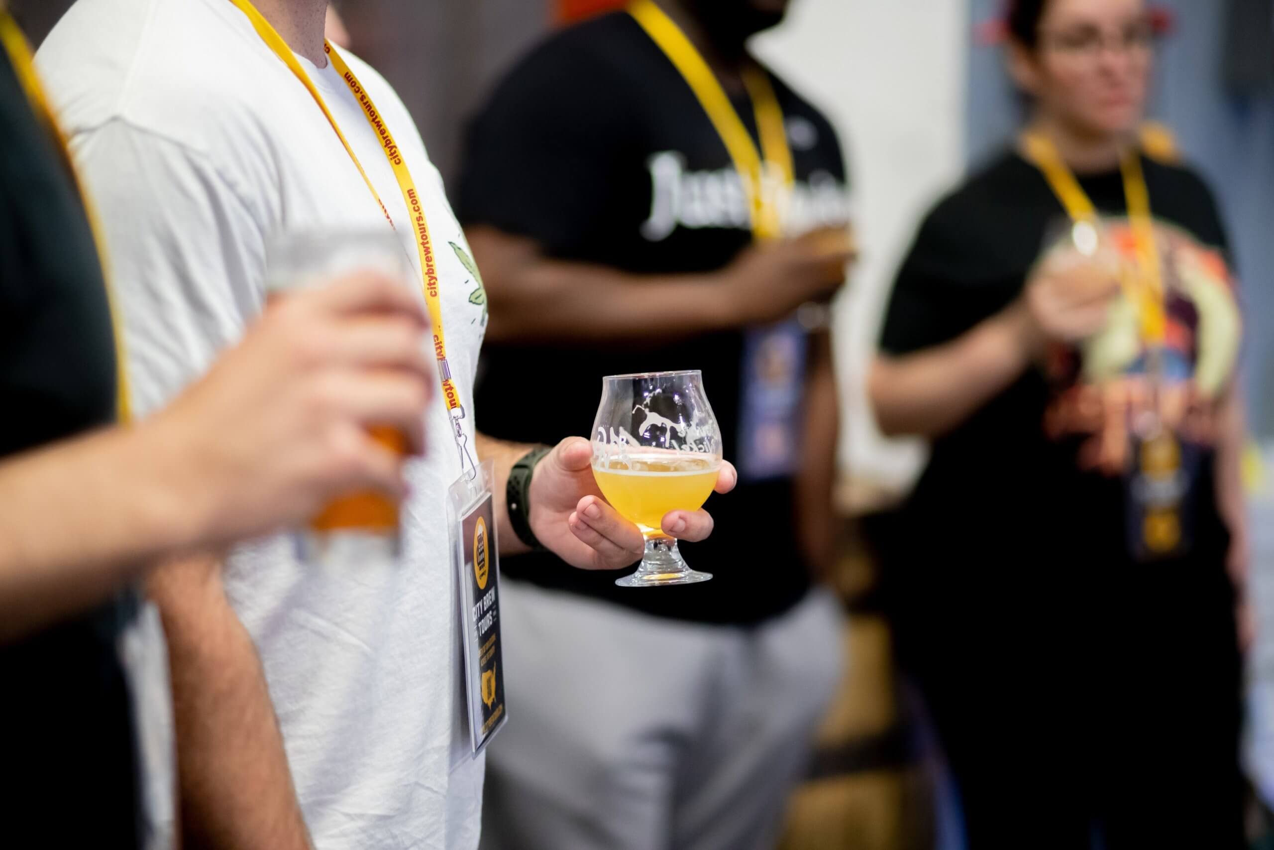 Four individuals are shown from the shoulders down. They are holding glasses of beer in their hands. Each person is wearing a yellow lanyard with a badge attached.