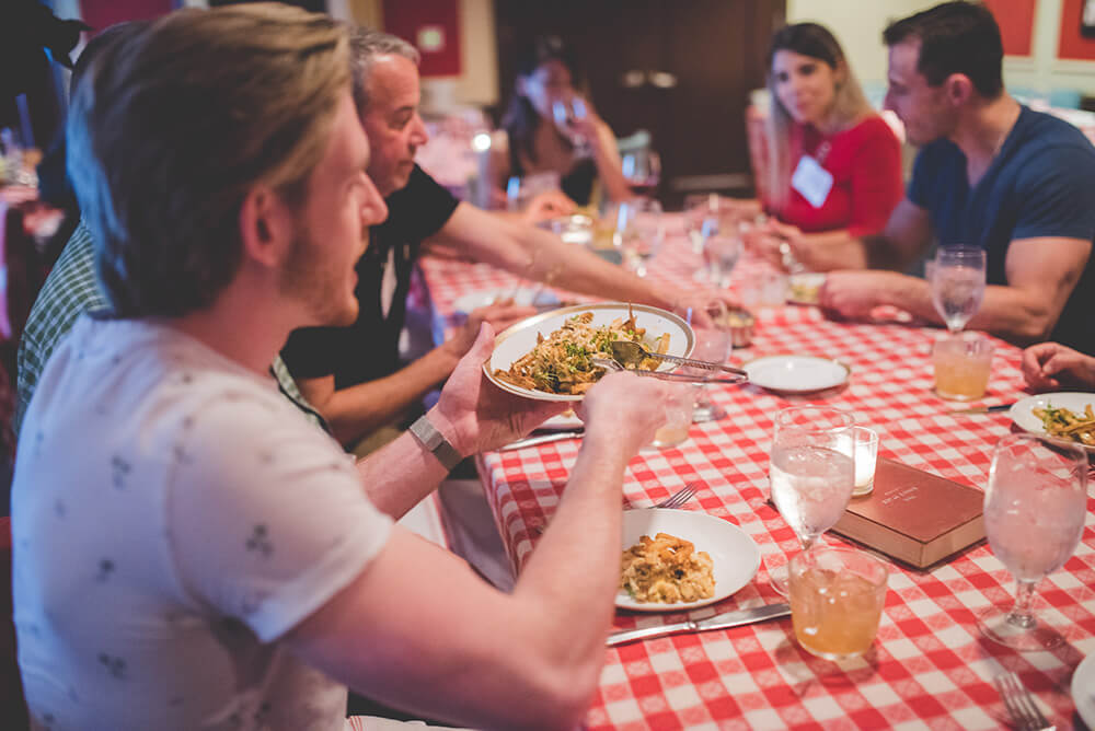 A man is shown is a white t-shirt, holding a plate of food in front of him. he sits at a table covered in a checkered red and white tablecloth. There are multiple other people sitting at the table.