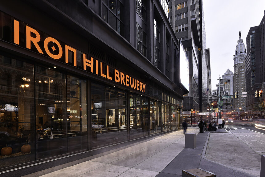 A large sign made of orange letters reads Iron Hill Brewery. The sign hangs across a building. The sidewalk in front of the brewery is clear. Off in the distance is Philadelphia City Hall.