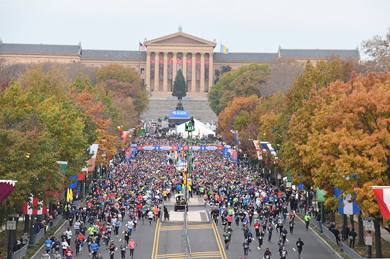 The Philadelphia Museum of Art is in the center. Runners are shown running away from the Philadelphia Museum of Art down the Benjamin Franklin Parkway. Colorful flags representing different countries wave in the wind, lining the parkway. The sky appears gray. The trees lining the road are green and orange, seasonal for fall.