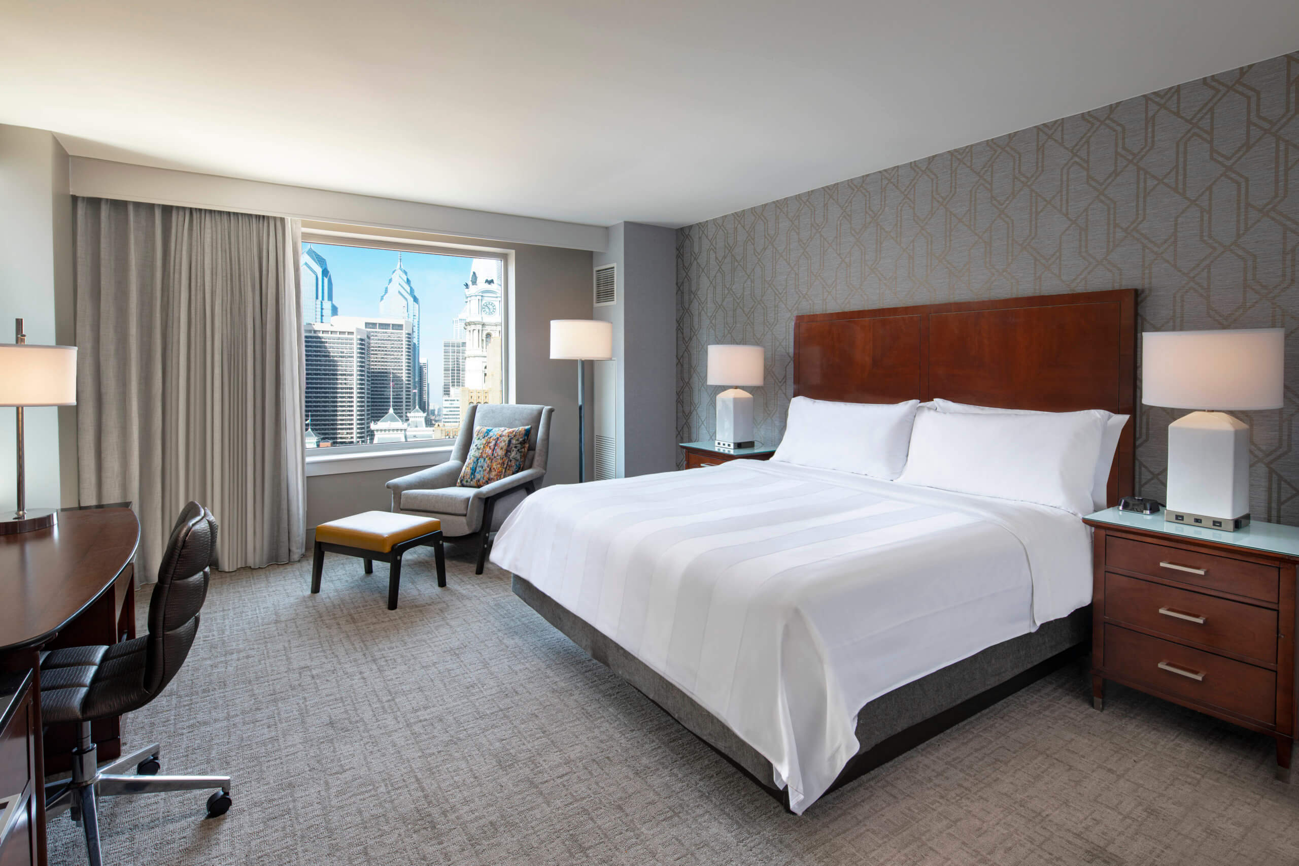 A desk and chair are off to the left. A lounge chair and ottoman are in the center of the frame in front of a window letting in natural light. A white bed with a wooden headboard is slightly off center with a nightstand next to it.