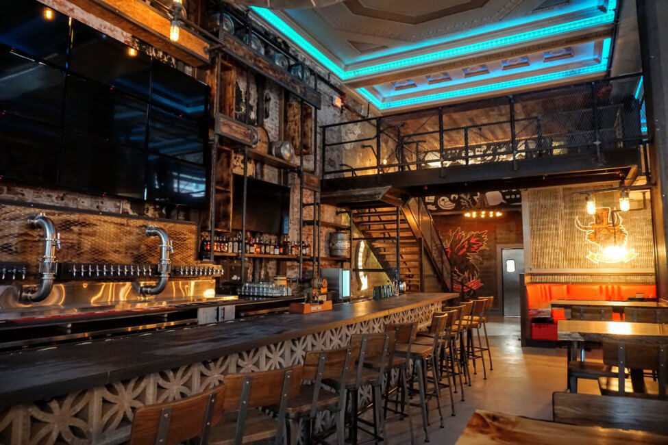 A large TV on a wall to the left hangs over a large bar. There are several bar stools pushed up against the bar. There is a set of stairs in the far left leading up to a balcony. The ceiling is glowing with blue lights. The overall feel of the bar is rustic with a lot of brown and metal accents throughout.