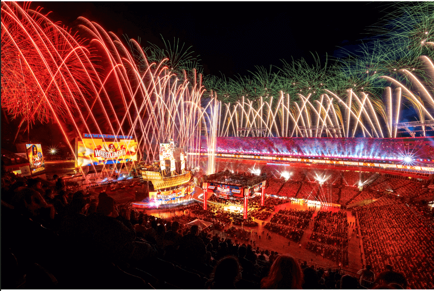 A stadium lights up as fireworks shoot into the sky. The main colors are red, yellow, and orange. Massive screens read WrestleMania. The arena is in the center, surrounded by bright lights as fans cheer from their seats.