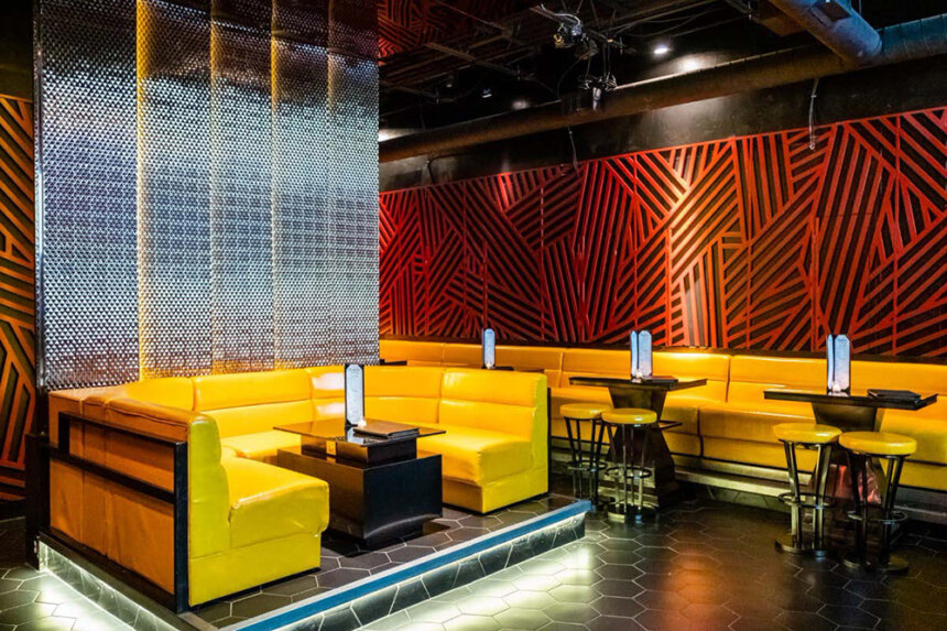 A bright yellow couch is shown in the center of the room with a black table in the middle. Along the wall on the right, there is a long yellow couch set up to serve as booths up against tables with two barstools by each one. The wall is decorated with a reddish orange design.