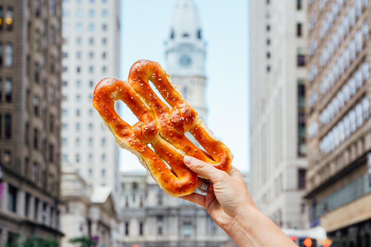 A hand is shown holding up a soft pretzel in front of Philadelphia City Hall.
