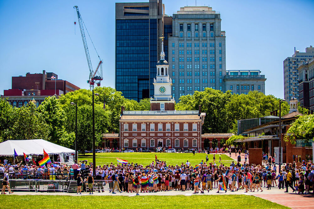 Independence Hall is in the background. A group of people are gathered in front. Some are shown with Pride rainbow flags. Others are dressed in bright colors. The grass is bright green. The sky overhead is a bright blue.