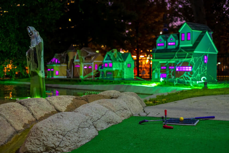 A miniature golf course is shown. There are lime green and purple lights lighting up the course. There are spooky, Halloween decor spread throughout. The small houses are covered in cobwebs.