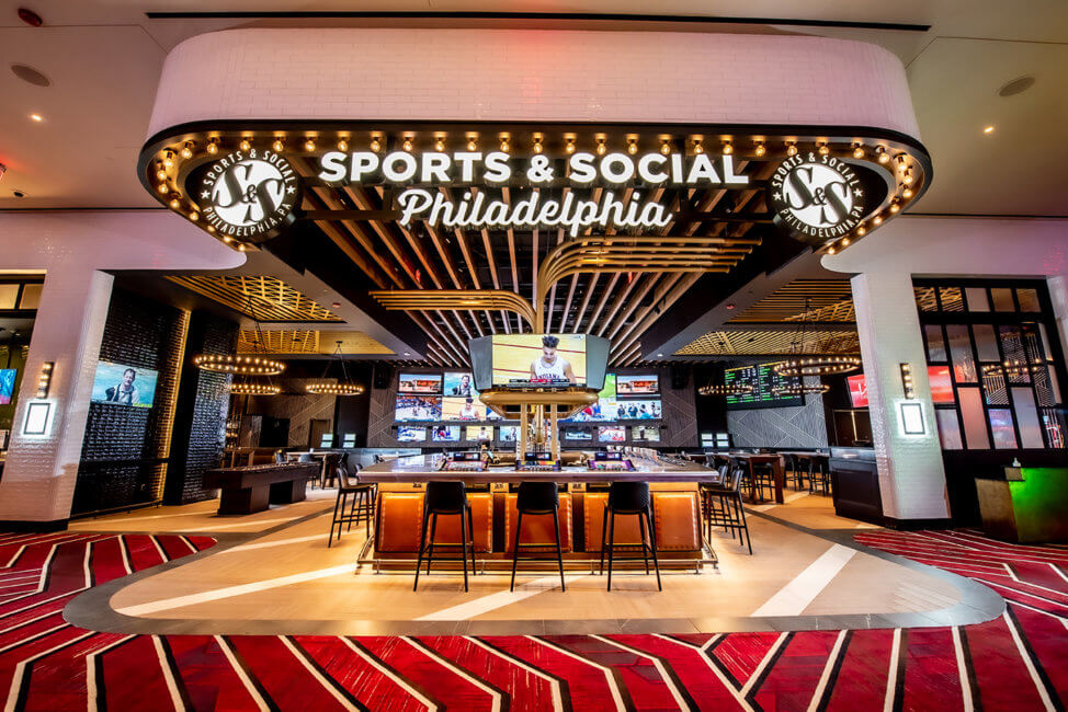 The carpet is a bright red with gold and black lines. A large space is shown with a sign reading Sports & Social Philadelphia. There are gold lights throughout the space. A TV hangs above a bar where there are barstools around either side. Beyond the bar in the front, there are several more screens with various sporting events being shown.