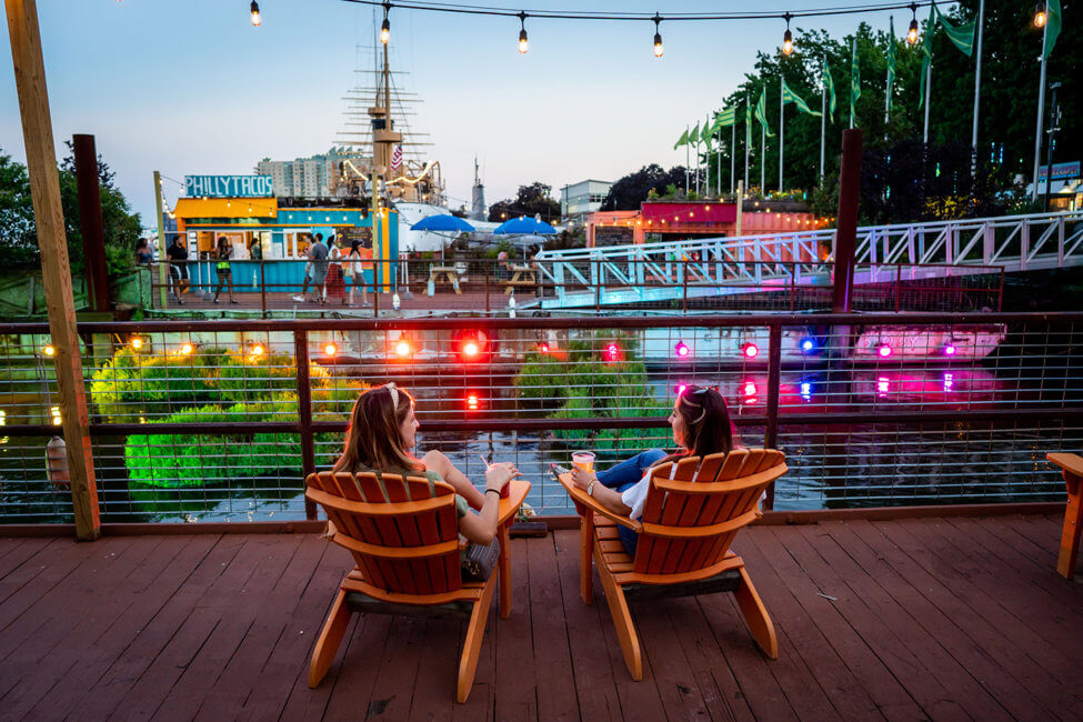 Two young women are shown smiling and holding drinks in their hands while sitting in orange Adirondack chairs on a boardwalk overlooking water. The water below them is lit up by colorful lights. Across the water, there is another area where multiple people are shown in front of a bright yellow and blue hut that has a sign that reads Philly Tacos.