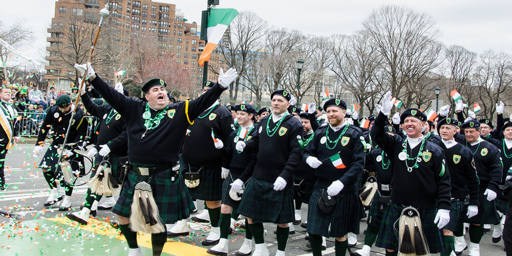 A large group of men dressed in black and green march down the street in celebration of Saint Patrick's Day. One man off to the left appears to lead the group. He has his arms stretched out and his hands in the air. All of the men are wearing hats to match their outfits. Green, orange, and white confetti has been tossed in the air.