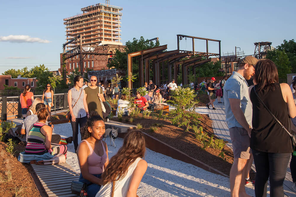 A bunch of people gather in an industrial park with beds of flowers and greenery.