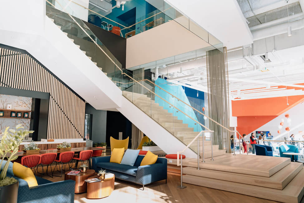 A bright open space is shown. There are colorful chairs, couches, pillows, and walls throughout. There is a grand staircase leading up to the upper floor. Off to the right, in front of an orange wall, there are multiple people shown standing around. To the left, two blue couches are shown with two yellow pillows on either side of a light blue pillow in the center. There are five orange chairs pushed up against a long table. Two centerpieces with plants are on the table.