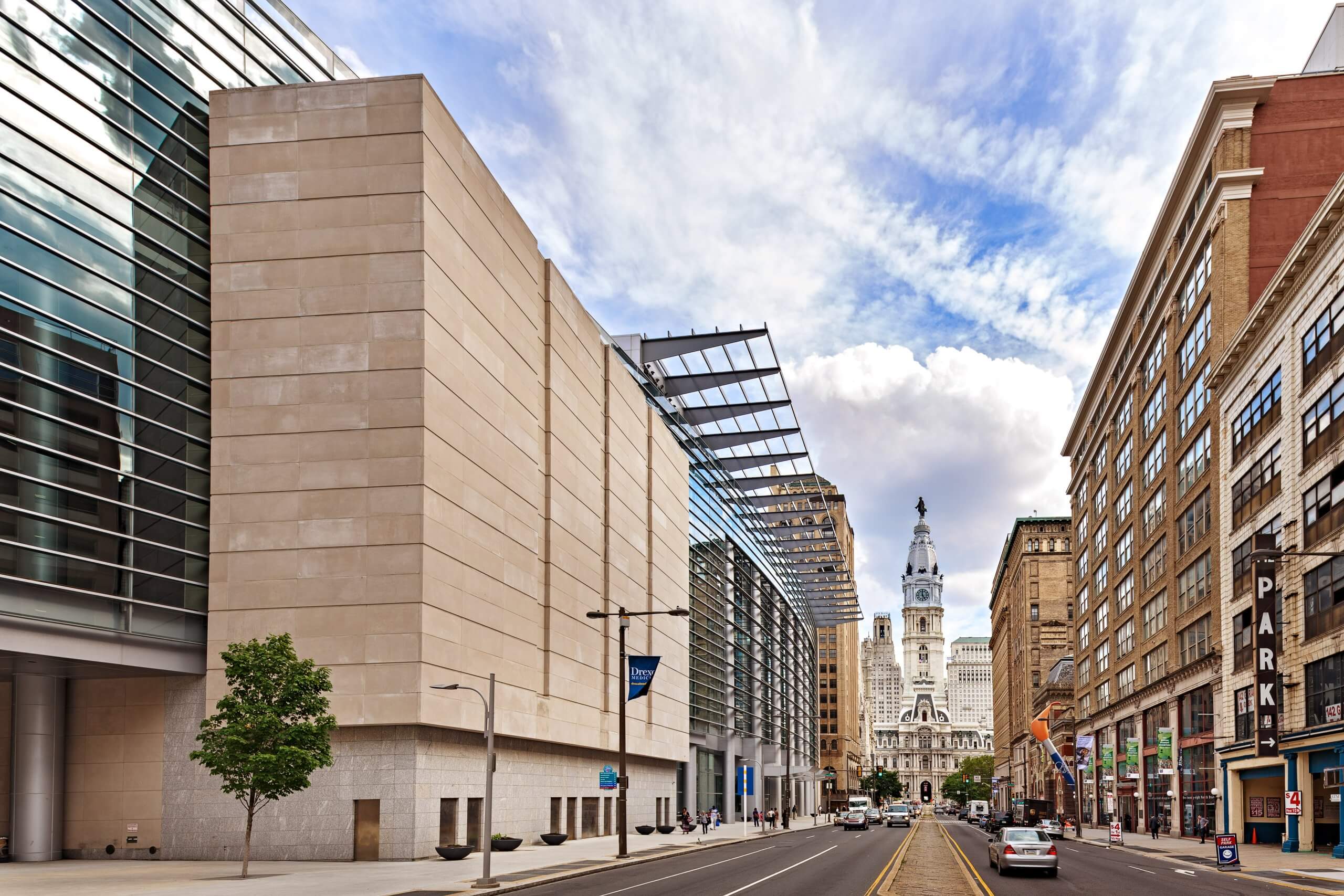 Outside of Pennsylvania Convention Center, a tree is in the bottom left hand corner of the frame, street is shown with a car driving toward City hall in the distance