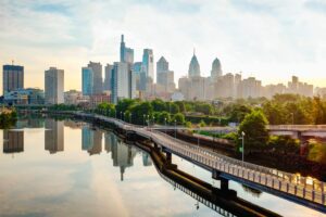 The sky is full of white clouds with parts of it blue. There is the Philadelphia skyline shown. There is a pedestrian bridge. Green trees are off to the right of it.