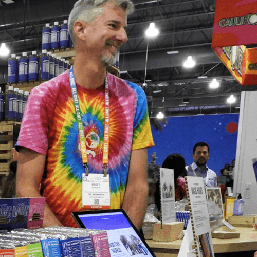 A man wearing a tie dyed t-shirt stands smiling looking off to the side. There are mint products on the table in front of him.