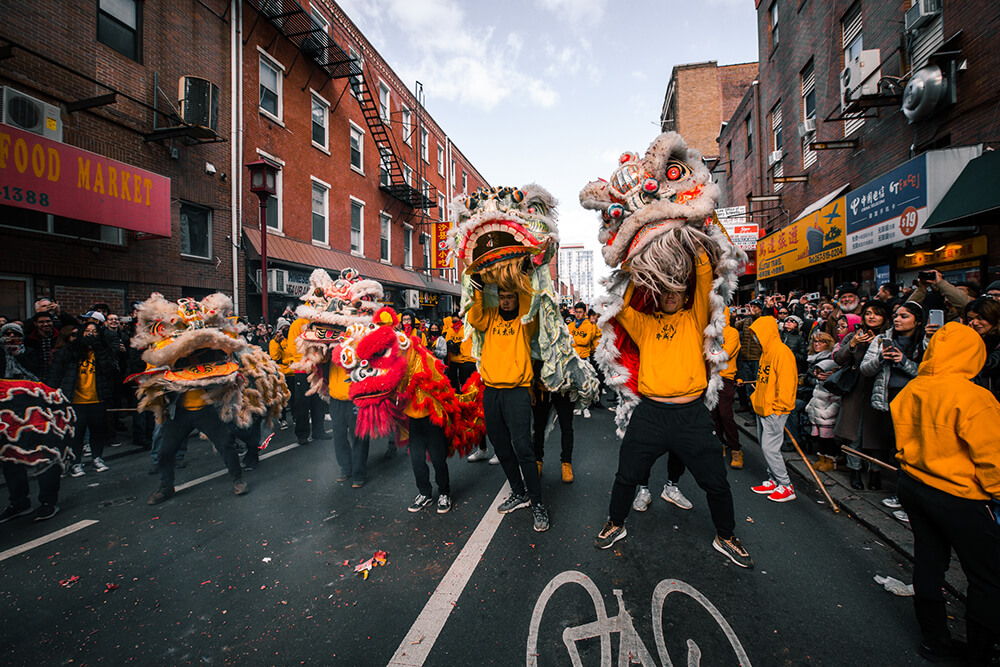 Multiple performers wearing yellow sweatshirts are holding up dragon heads as part of a costume as they dance in the street during a Lunar New Year parade.