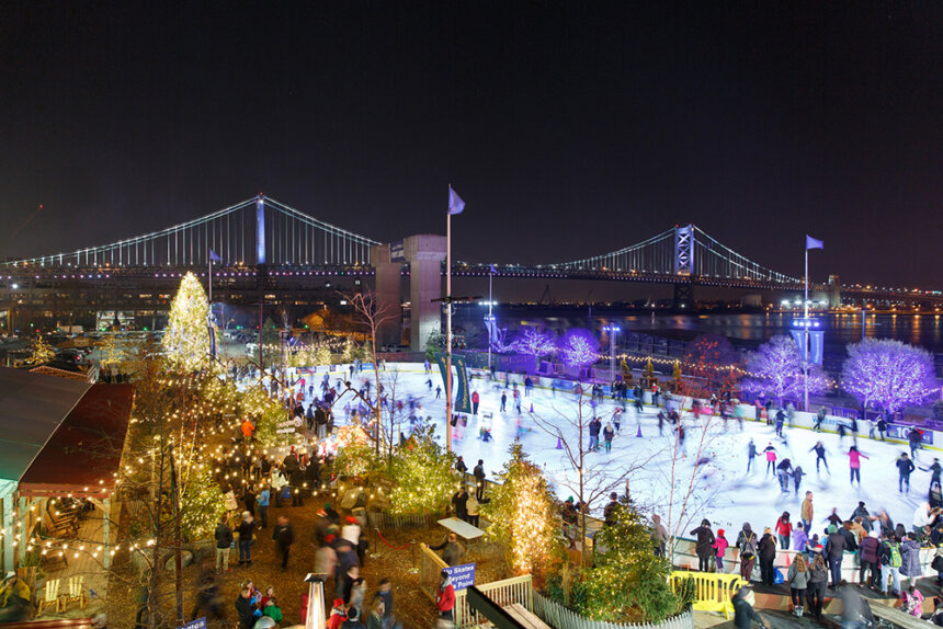 It is nighttime. A large outdoor ice rink is shown. There are multiple people ice skating. A bridge is shown off in the distance.