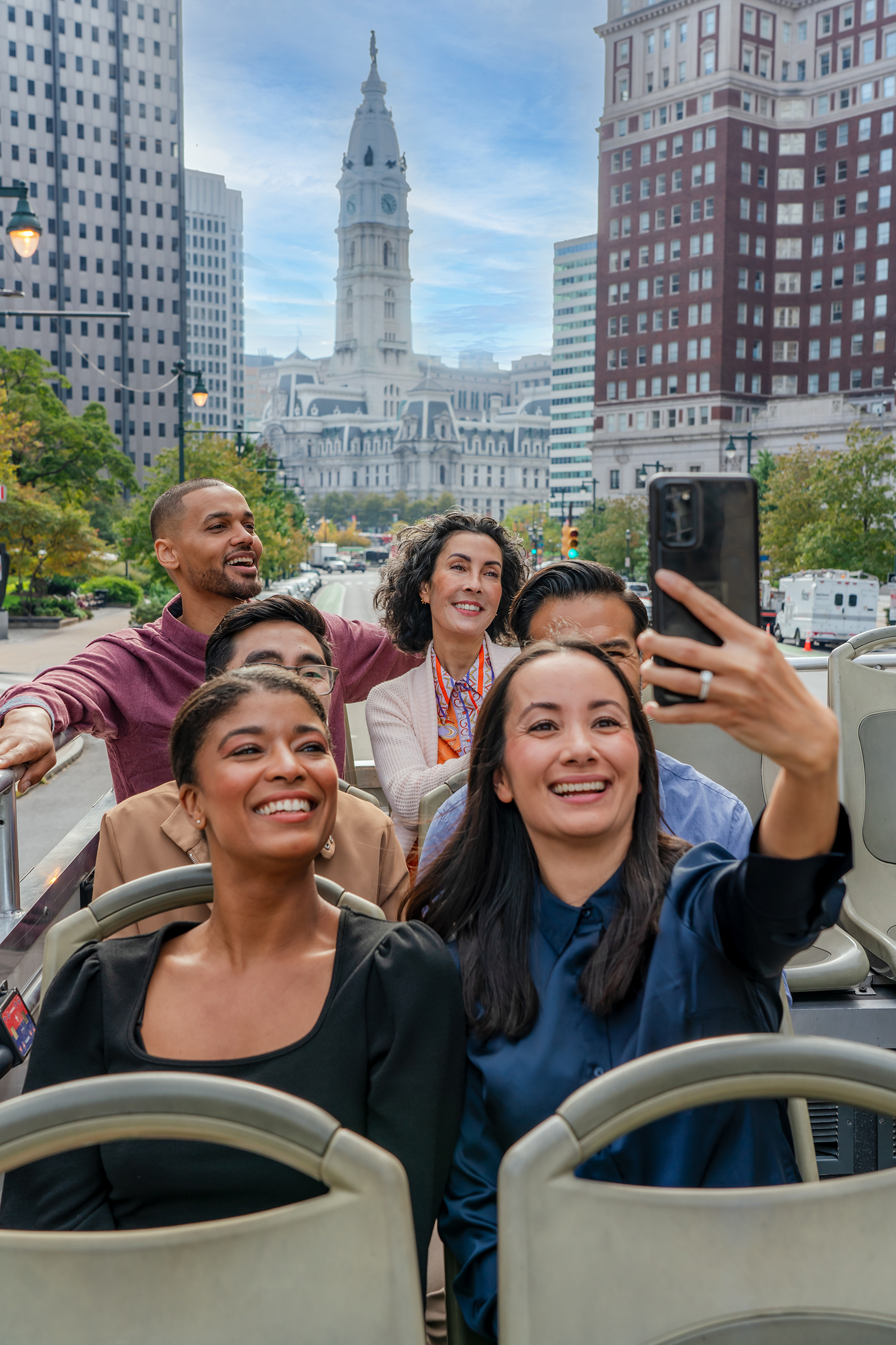 A group of people on a tour bus take a selfie with Philadelphia's City Hall in the background.