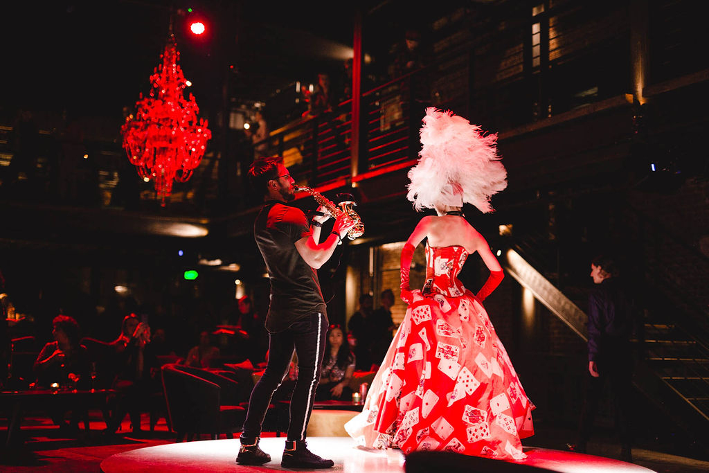A woman in a red dress stands off to the right. A man is to her left, he is playing a saxophone, a gold musical instrument. He is dressed in all black and is wearing sneakers. The room they are in is dark, but a chandelier hangs from the ceiling and appears to be glowing red.