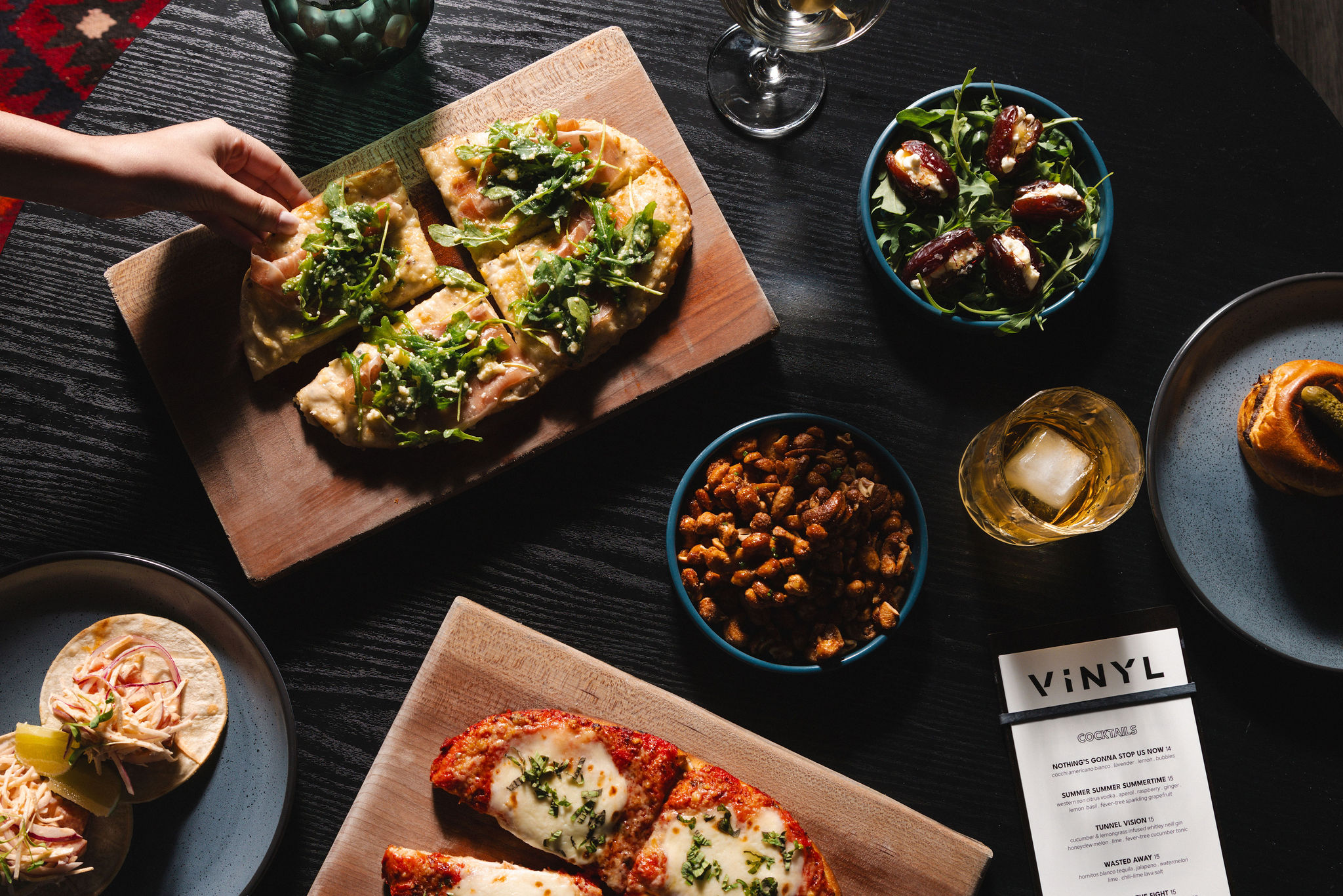 An above the table angle of food offerings at Vinyl, including a prosciutto and arugula pizza, chickpea salad, and tacos.