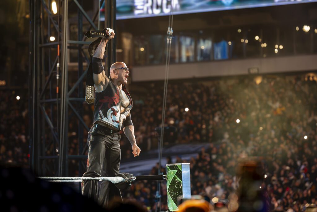 The Rock holds a microphone over his head to the crowd on stage at WrestleMania XL Night 1 at Lincoln Financial Field.