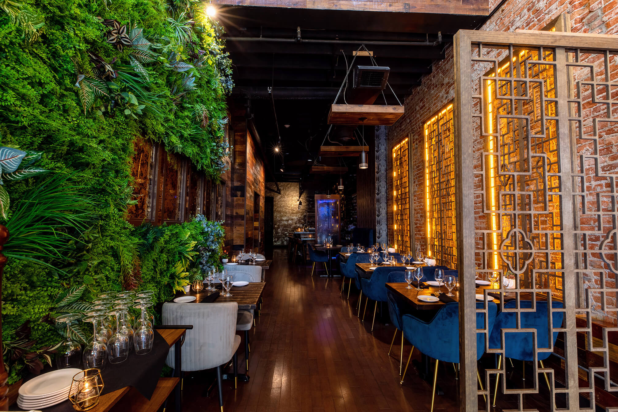 The inside of a restaurant is shown. There is greenery and plants on the wall to the left. There are wine glasses placed upside down on a table in front of the greenery. There are tables lined up in the space with gray chairs pushed up against the tables of the left and blue chairs pushed up against the tables on the right. There are plates, silverware, and glasses on each table. There is glowing gold light shining through the wall on the right. The wooden floors are a dark brown.