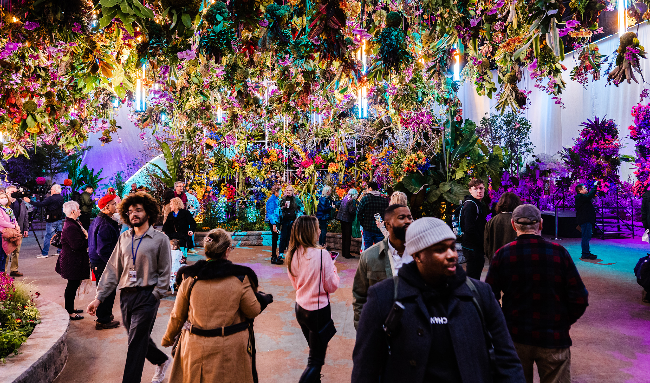 Bright, colorful floral arrangements hang from the ceiling. Flower Show attendees walk underneath, marveling at the floral displays surrounding them. A large floral display is in the center with a crowd of people admiring it.