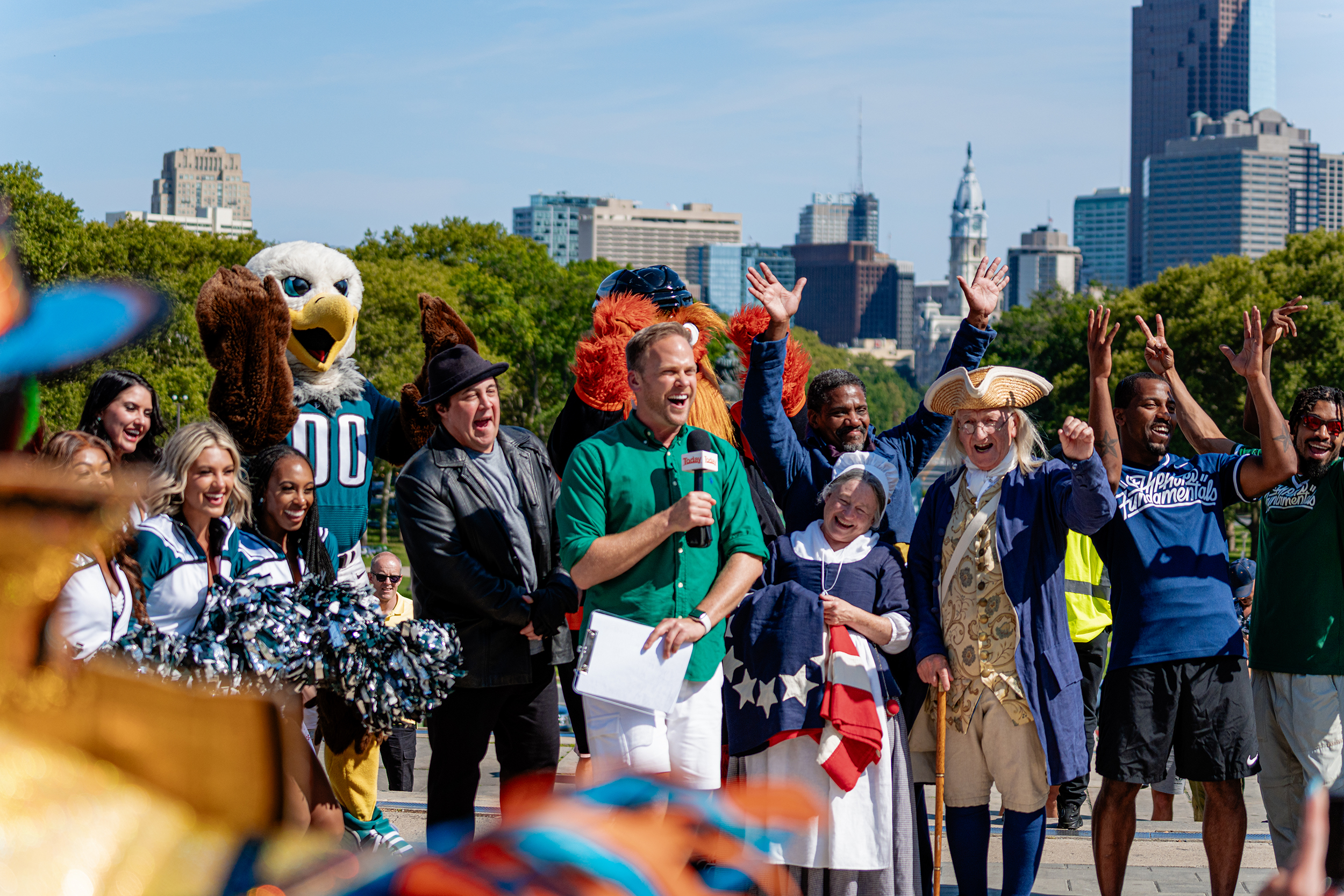 A news reporter is surrounded with Philadelphia's mascots and the city skyline in the background.