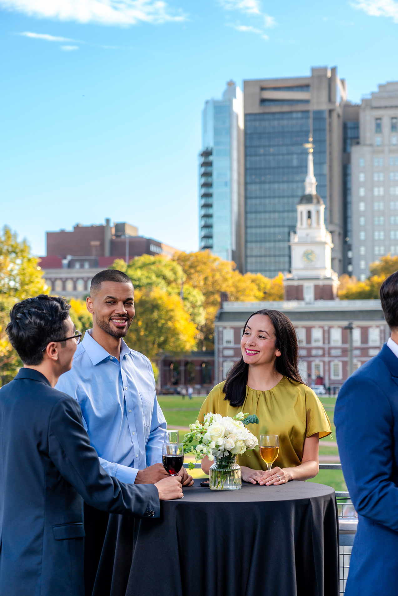 Three people speak with each other around a table. Independence Hall is in the background.