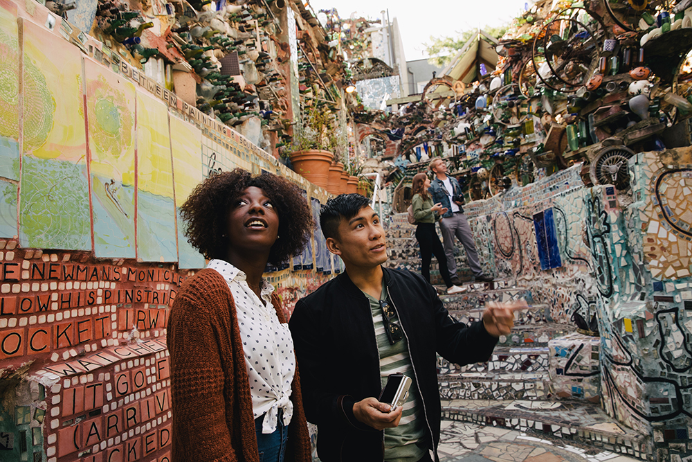 A woman and a man stand admiring an outdoor mosaic garden. there are colorful tiles and broken glass bottles held together by colorful grout.