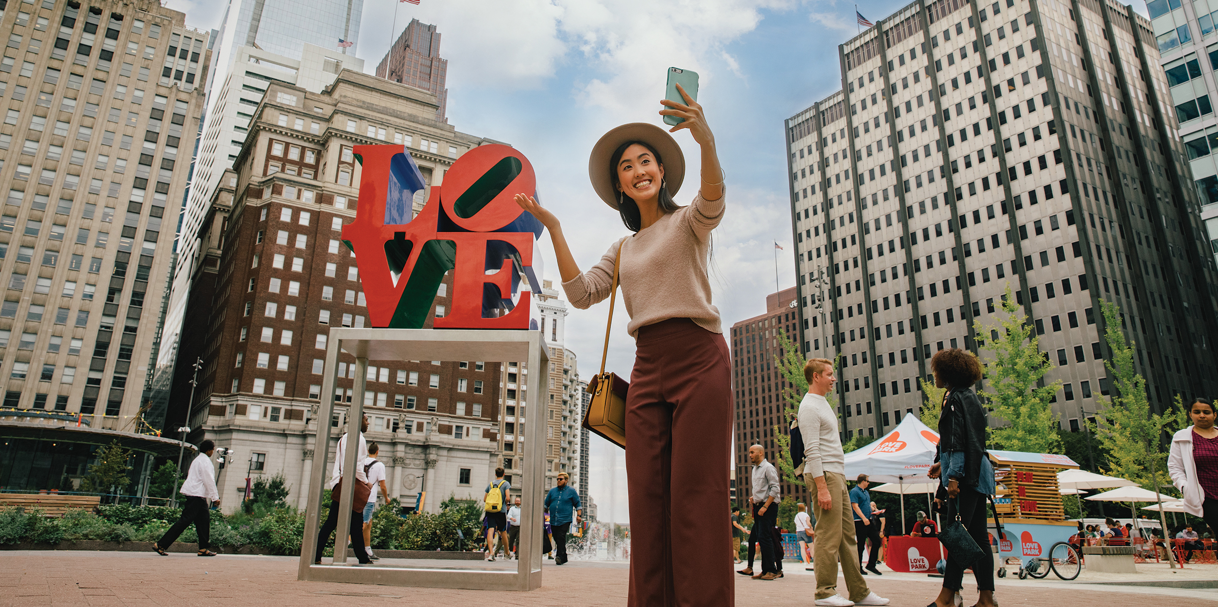 A woman wearing auburn pants, a beige sweater, and a tan hat poses for a selfie in front of the iconic red LOVE sculpture in LOVE Park. People throughout the space are shown behind her.