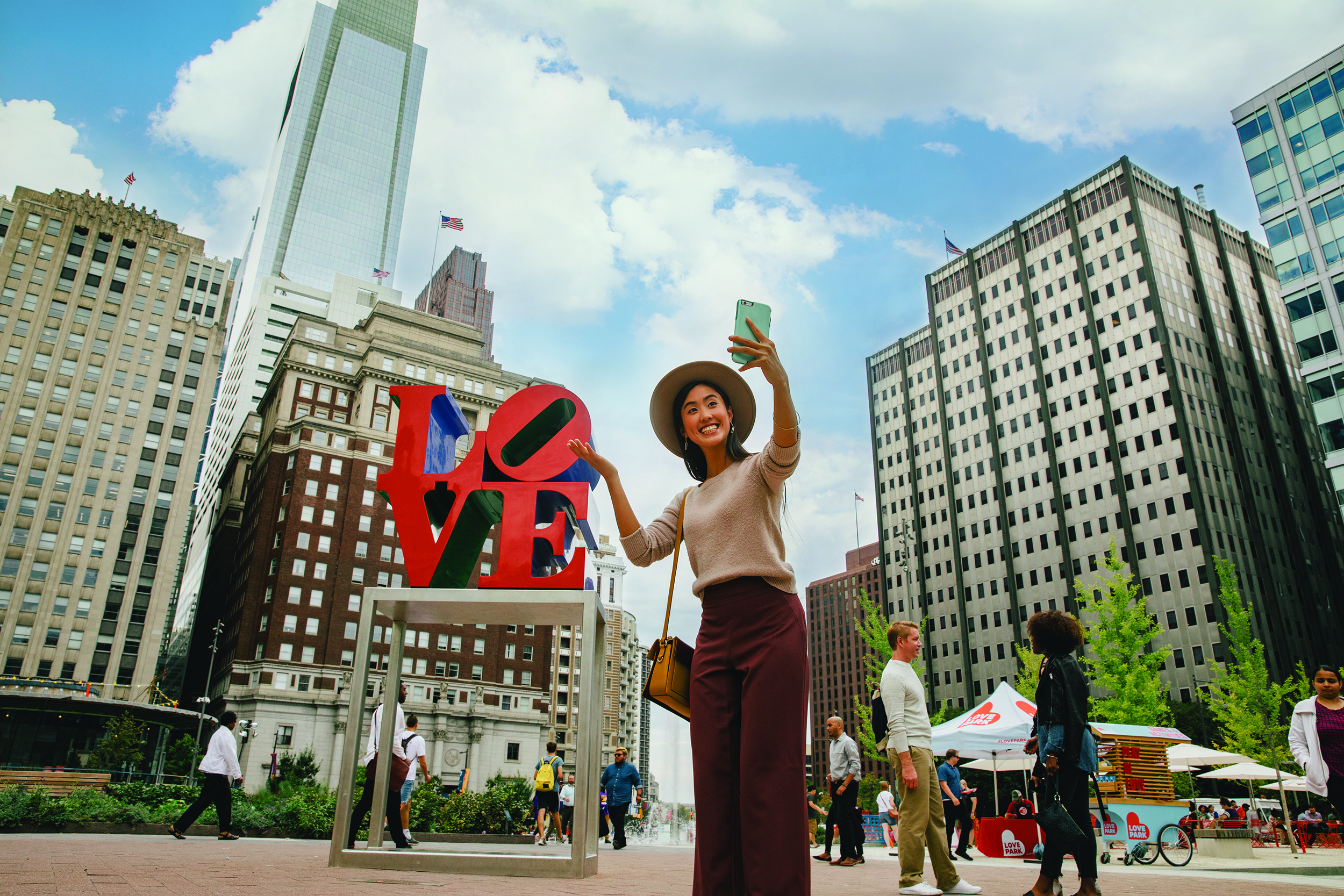 A woman wearing auburn pants, a beige sweater, and a tan hat poses for a selfie in front of the iconic red LOVE sculpture in LOVE Park. People throughout the space are shown behind her.