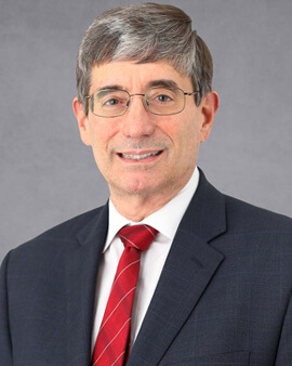 A man with gray hair who is wearing glasses, a black suit jacket, white shirt, and red tie is shown smiling for the camera.