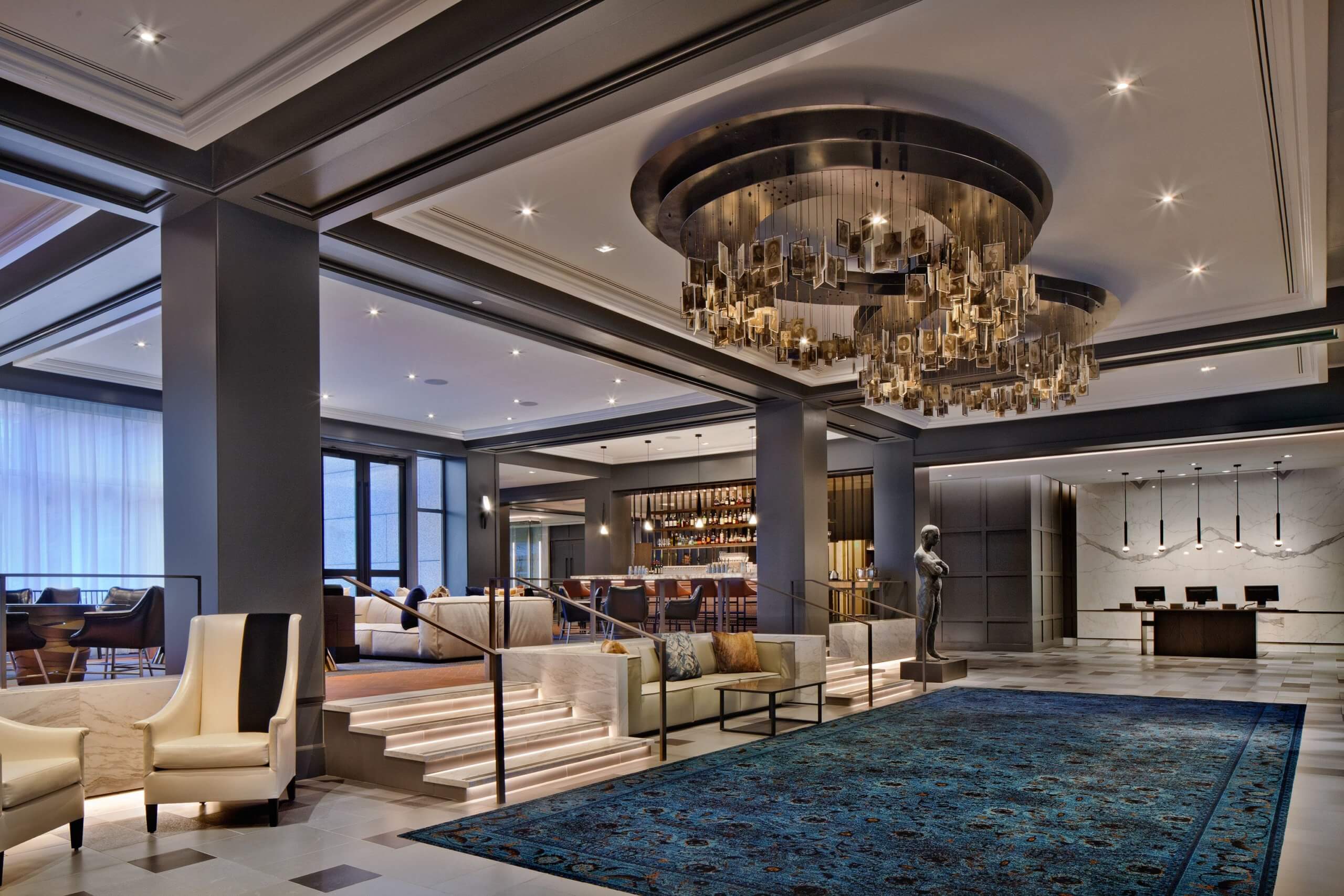 A hotel lobby is shown. There is a large chandelier made of photos hanging from the ceiling. There is a blue carpet underneath. There are a few steps off to the left leading up to another level.