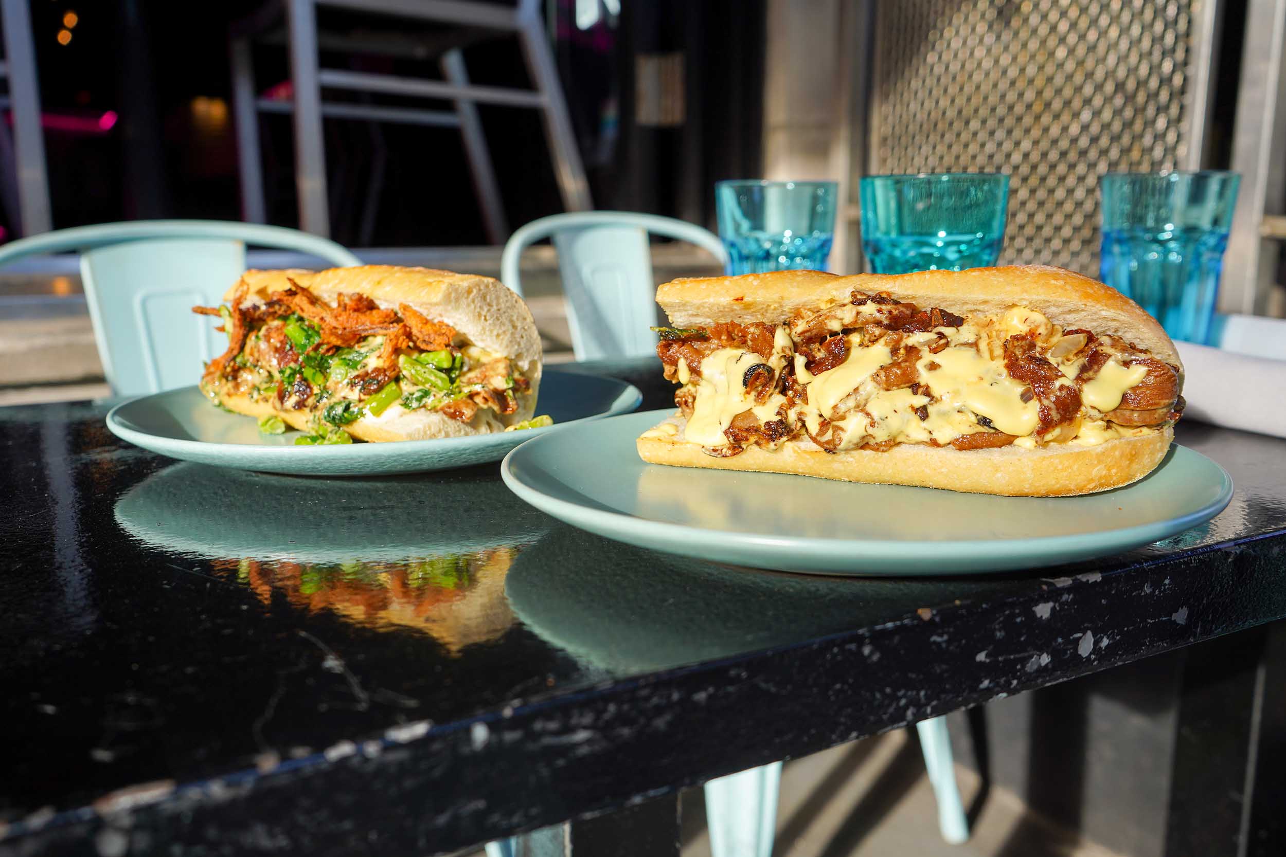 Two vegan cheesesteaks sit on a table in the sun.