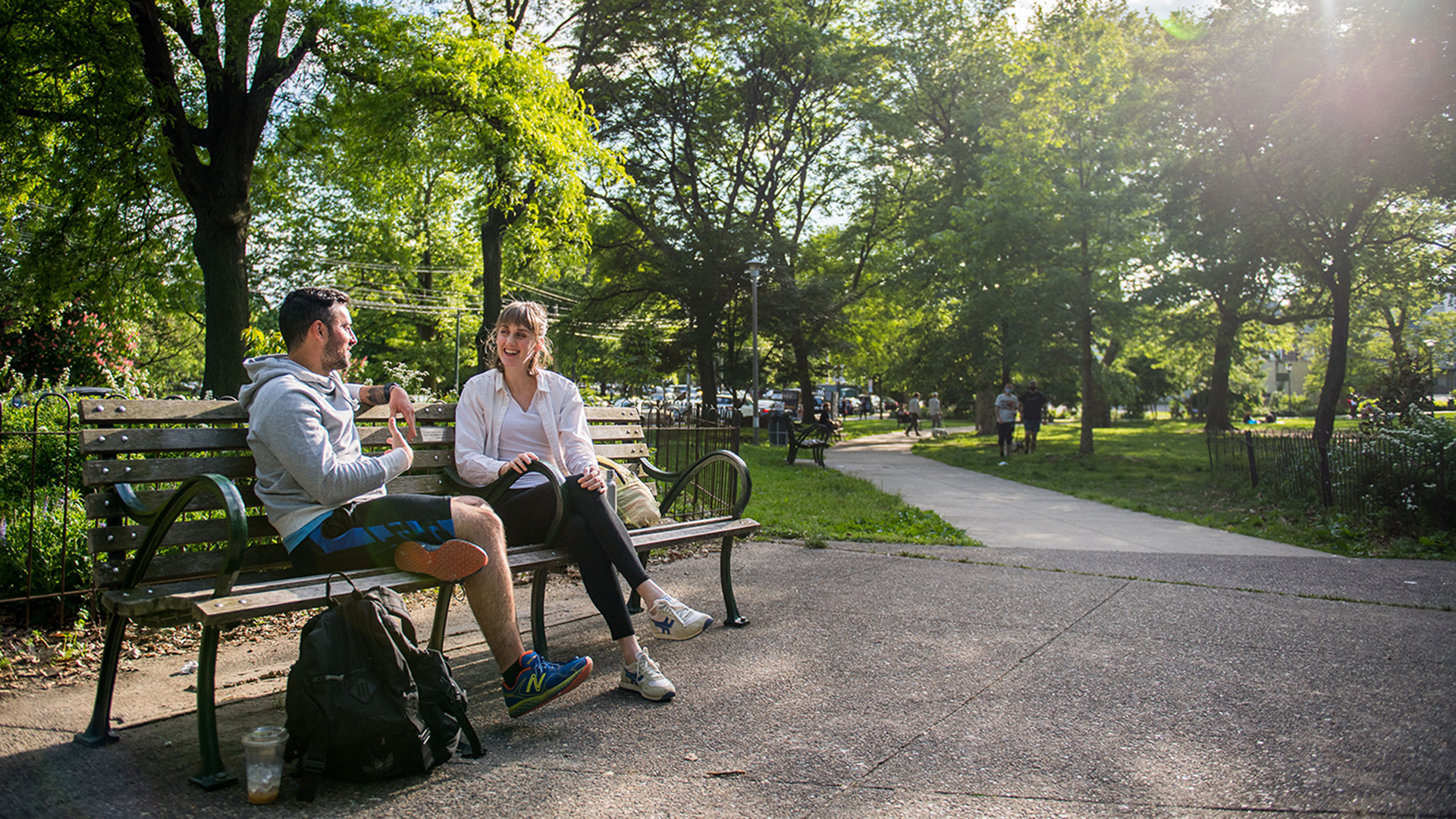Two people sit on an bench, talking, in front of a lush, green park.
