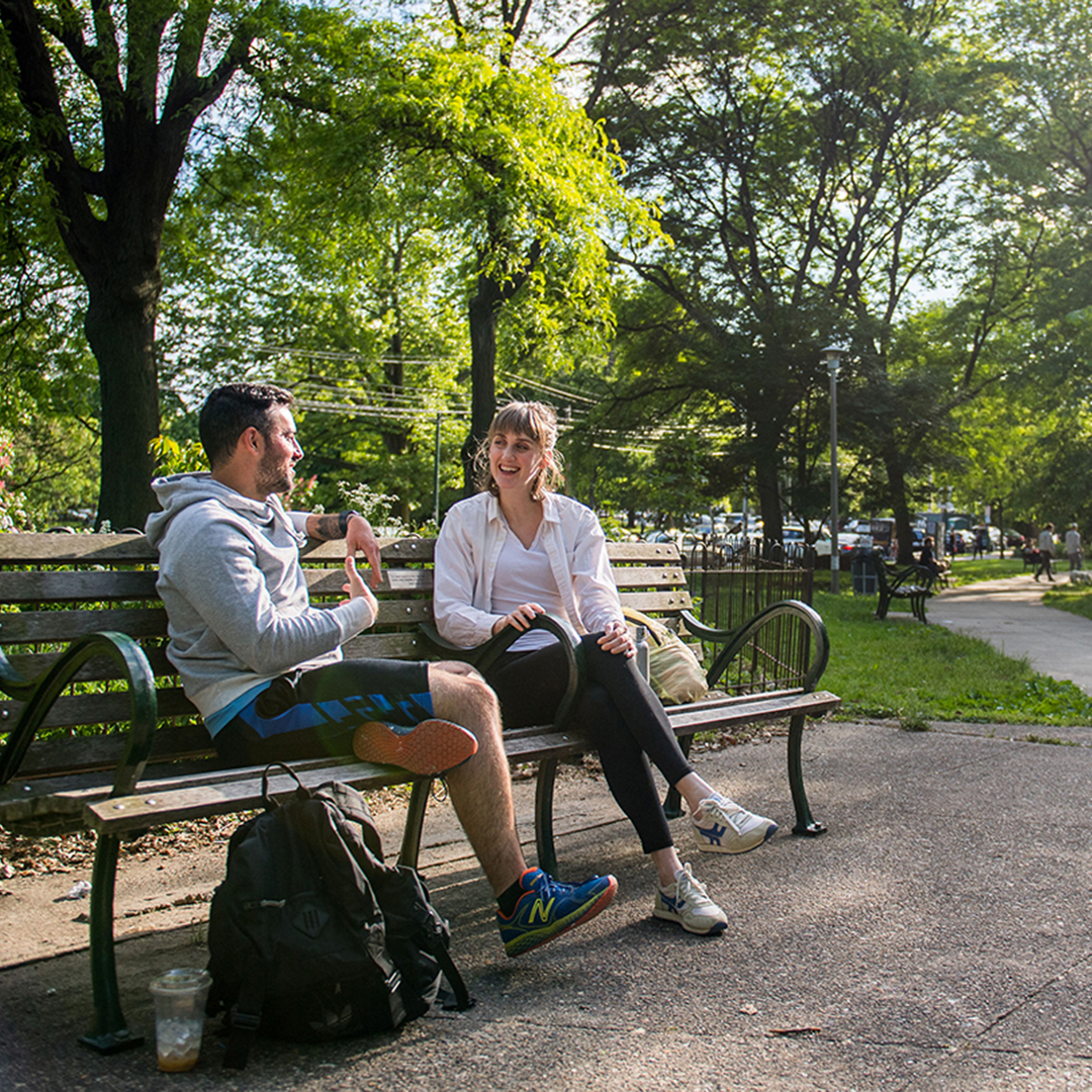Two people sit on an bench, talking, in front of a lush, green park.