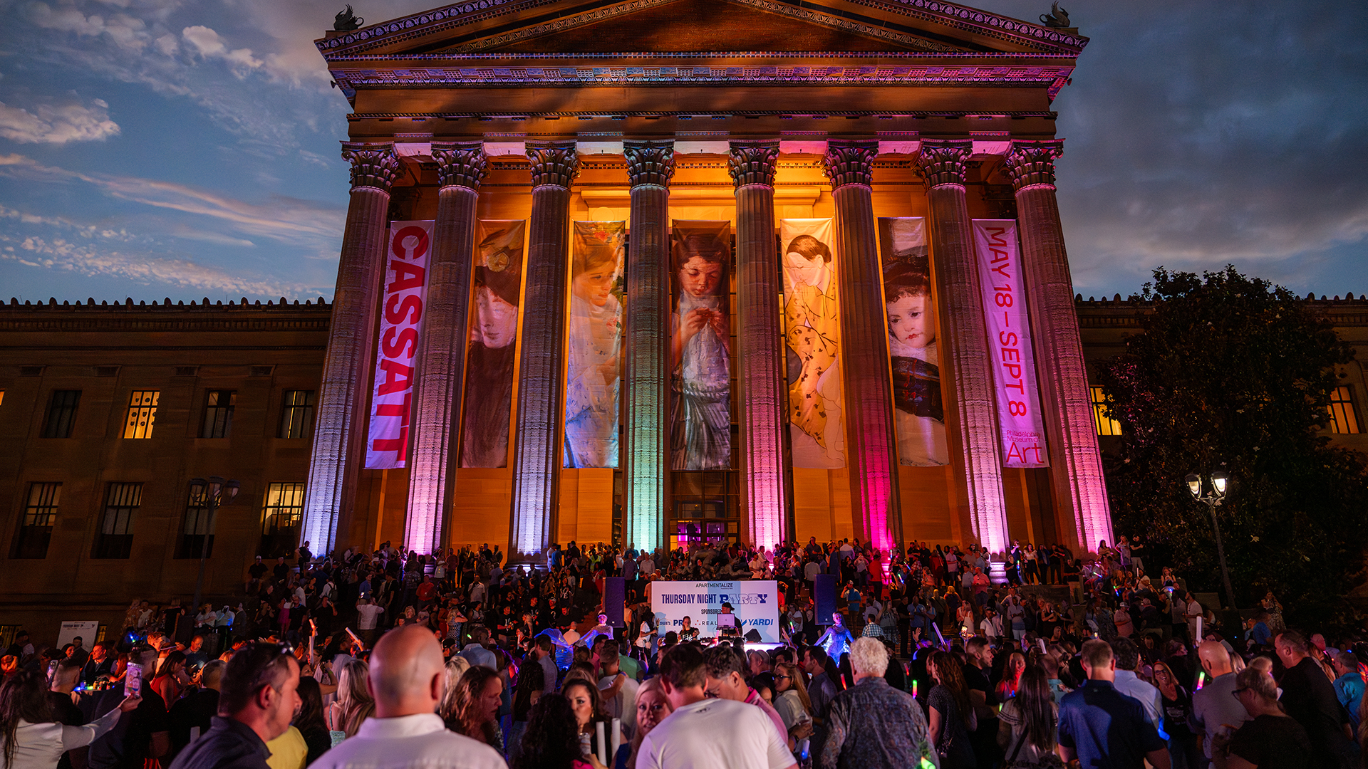 A huge gathering outside of the Philadelphia Museum of Art. The Museum's columns are lit up with colorful lights.