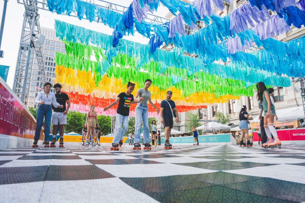 Roller-skaters undeneath a rainbow banner, skate on the Dilworth Park roller rink.